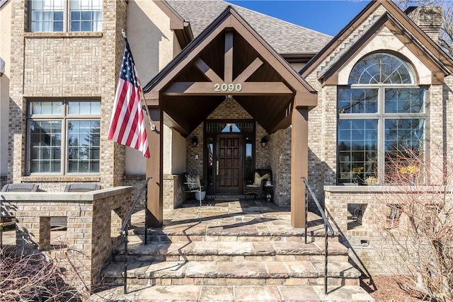 view of exterior entry with stucco siding, brick siding, and roof with shingles