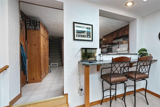 kitchen with tile patterned floors, under cabinet range hood, dark countertops, baseboards, and a textured wall