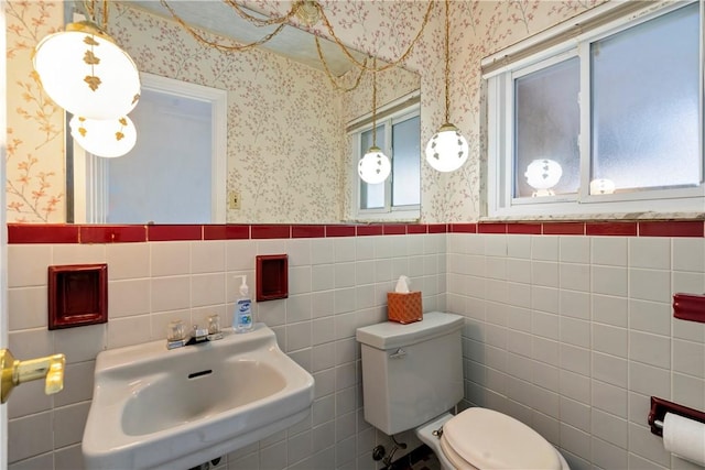 bathroom featuring wallpapered walls, a sink, wainscoting, tile walls, and toilet