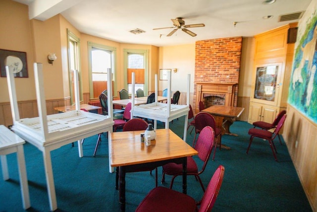 carpeted dining room with a wainscoted wall, a fireplace, a ceiling fan, and visible vents