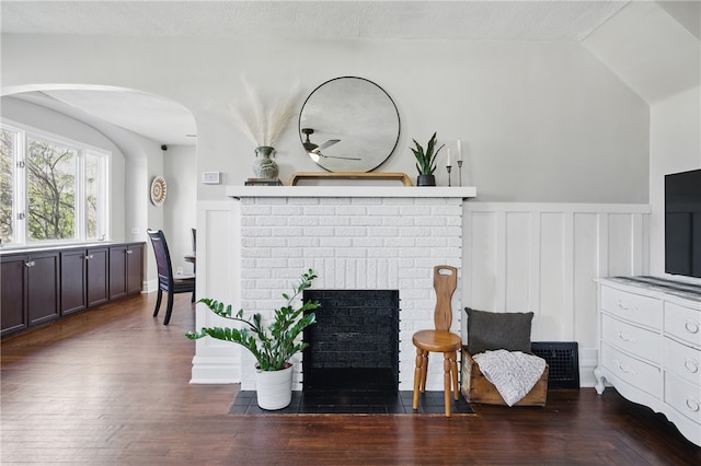 living area featuring a wainscoted wall, dark wood finished floors, lofted ceiling, a textured ceiling, and a brick fireplace