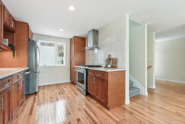 kitchen with light wood finished floors, decorative backsplash, brown cabinets, appliances with stainless steel finishes, and wall chimney exhaust hood