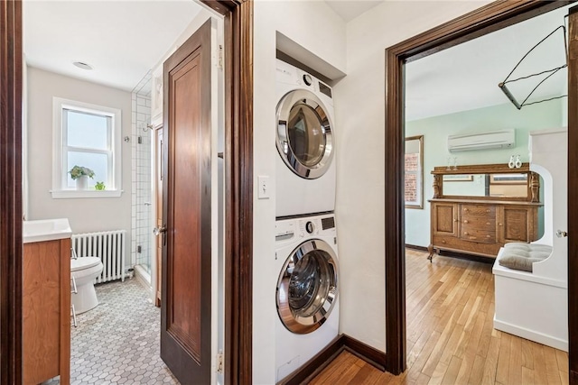 clothes washing area with an AC wall unit, radiator heating unit, stacked washing maching and dryer, light wood finished floors, and laundry area