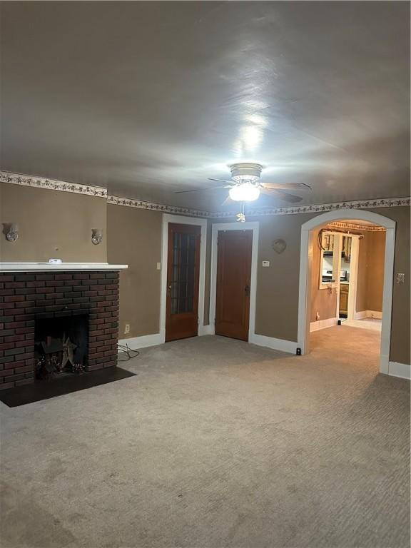 unfurnished living room featuring a brick fireplace, baseboards, carpet, arched walkways, and a ceiling fan