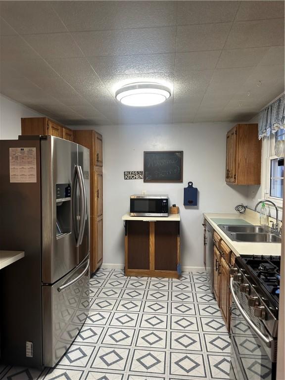 kitchen with a sink, appliances with stainless steel finishes, brown cabinetry, light countertops, and baseboards