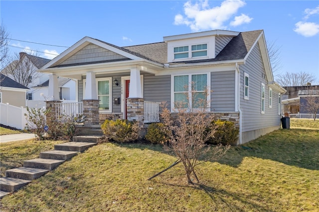 view of front of home with a front lawn, fence, roof with shingles, covered porch, and stone siding