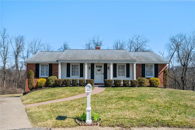 ranch-style home featuring a front lawn, covered porch, a shingled roof, brick siding, and a chimney