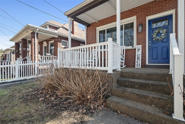 view of exterior entry featuring brick siding, covered porch, and fence