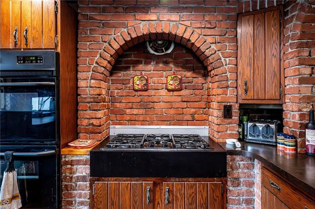 kitchen featuring dobule oven black, gas cooktop, dark countertops, brick wall, and brown cabinetry