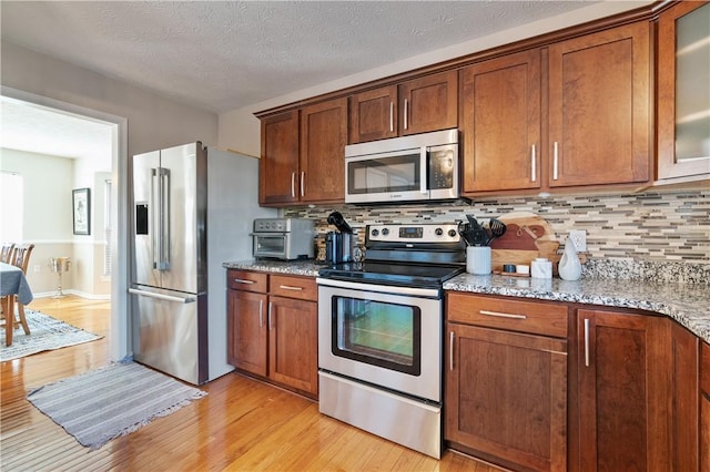 kitchen featuring light wood-type flooring, decorative backsplash, light stone counters, appliances with stainless steel finishes, and glass insert cabinets