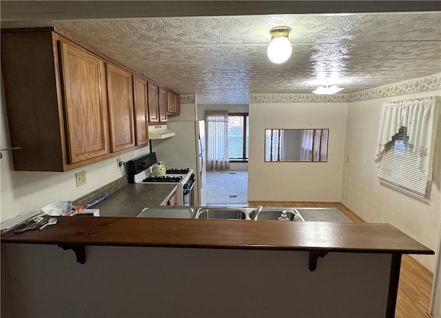 kitchen featuring white gas stove, under cabinet range hood, a textured ceiling, dark countertops, and brown cabinets