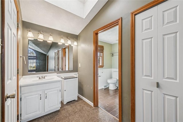 bathroom with lofted ceiling with skylight, a wainscoted wall, toilet, and vanity