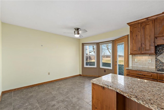 kitchen featuring brown cabinets, a ceiling fan, light stone counters, backsplash, and baseboards