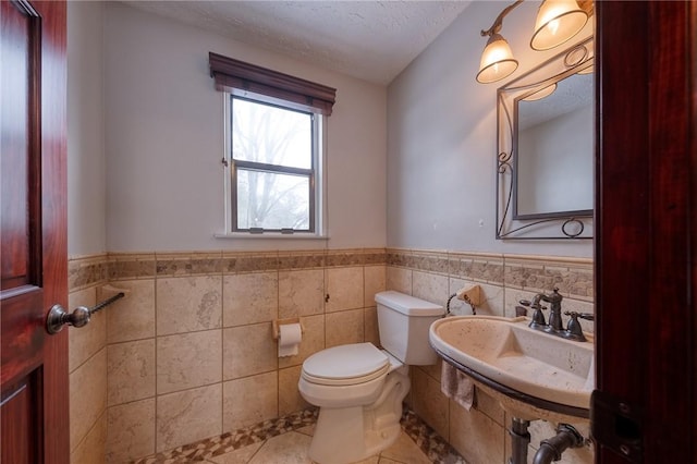 half bath with a wainscoted wall, toilet, tile patterned floors, a textured ceiling, and a sink