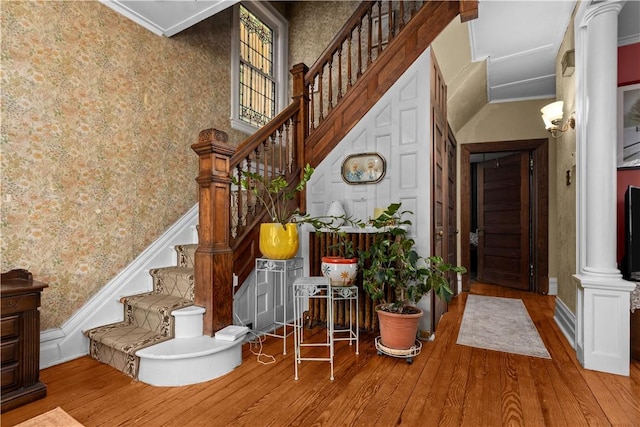 staircase featuring wood finished floors, ornate columns, wallpapered walls, a towering ceiling, and crown molding