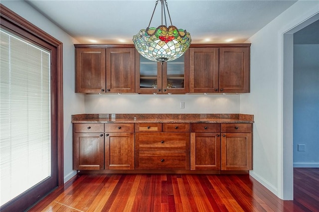 kitchen featuring dark wood-style floors, baseboards, dark stone counters, hanging light fixtures, and glass insert cabinets