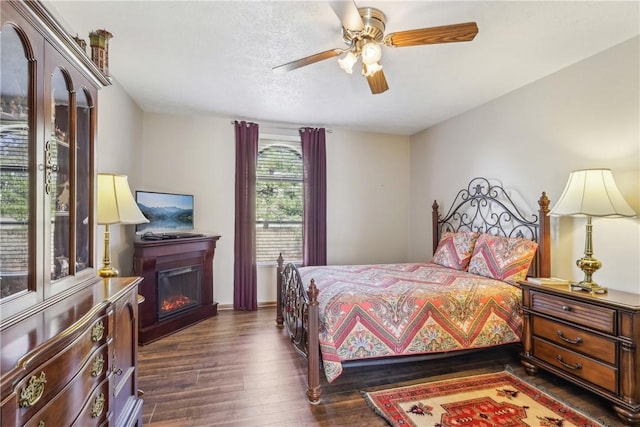 bedroom featuring a glass covered fireplace, dark wood-type flooring, and ceiling fan