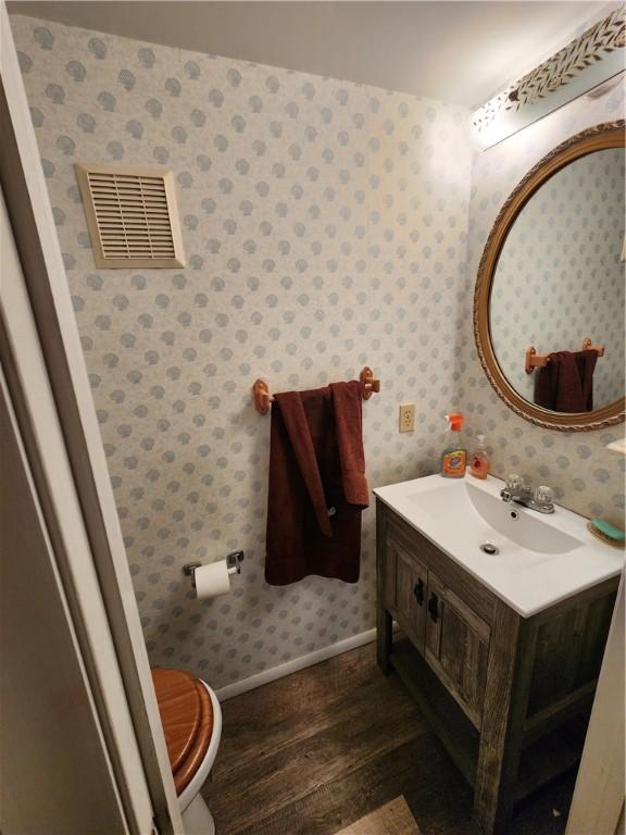 half bath with visible vents, wood finished floors, wallpapered walls, baseboards, and vanity