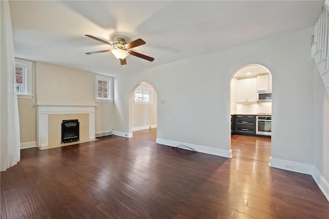 unfurnished living room featuring baseboards, arched walkways, ceiling fan, dark wood-type flooring, and a tiled fireplace