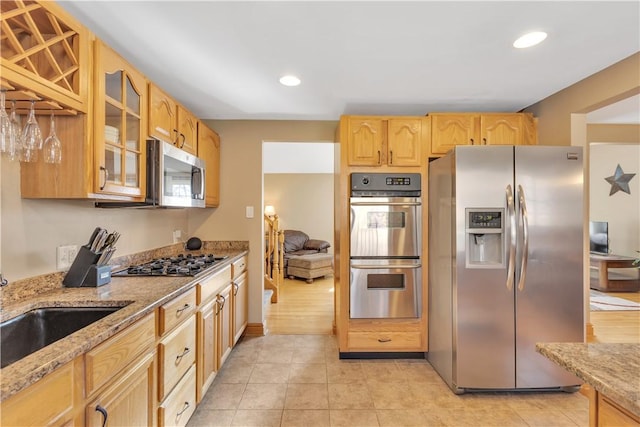 kitchen with glass insert cabinets, light stone countertops, appliances with stainless steel finishes, and light brown cabinetry