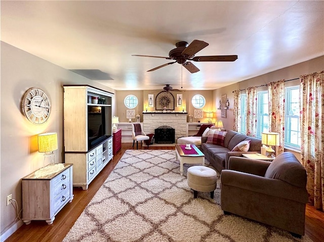 living area featuring a stone fireplace, baseboards, dark wood-type flooring, and a ceiling fan