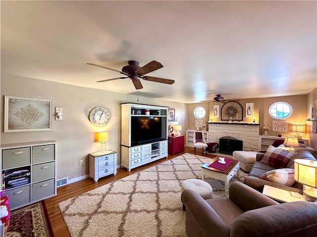 living area featuring visible vents, baseboards, a stone fireplace, wood finished floors, and a ceiling fan