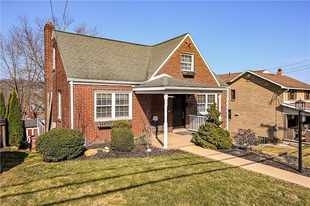 view of front of property featuring a front yard, roof with shingles, covered porch, a chimney, and brick siding