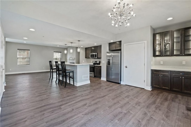 kitchen with dark wood-style floors, baseboards, an island with sink, dark brown cabinets, and appliances with stainless steel finishes