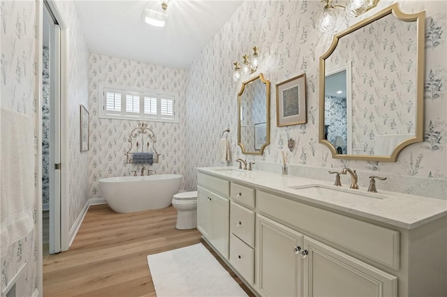 bathroom featuring wood finished floors, wallpapered walls, a soaking tub, double vanity, and a sink