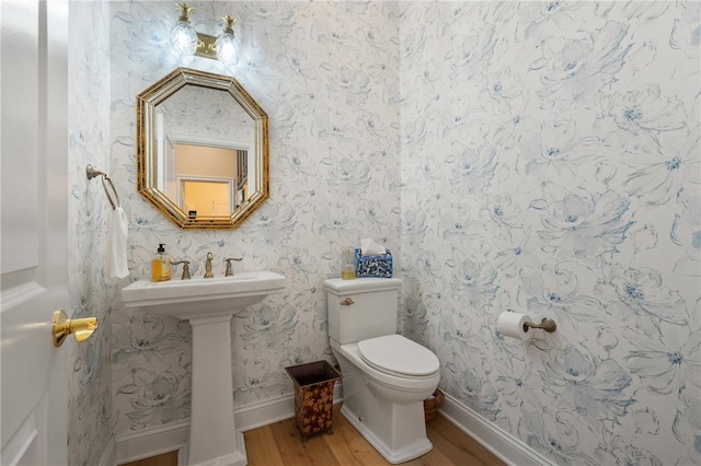 half bathroom featuring wood finished floors, baseboards, wallpapered walls, a sink, and toilet