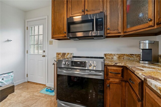 kitchen with glass insert cabinets, baseboards, light stone countertops, brown cabinets, and stainless steel appliances