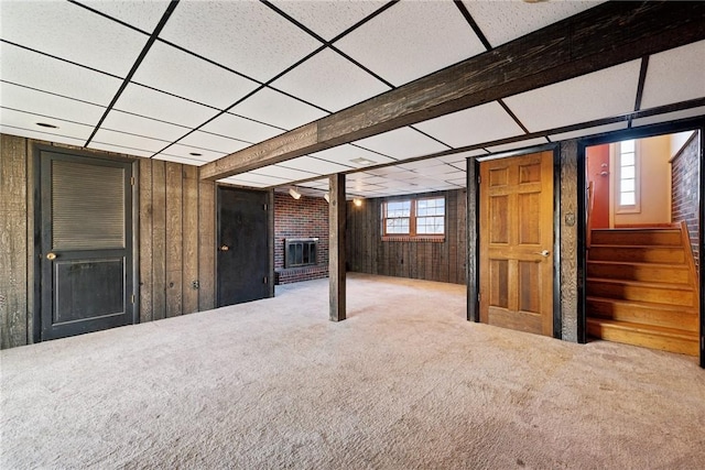 below grade area with a drop ceiling, carpet floors, wooden walls, and a brick fireplace