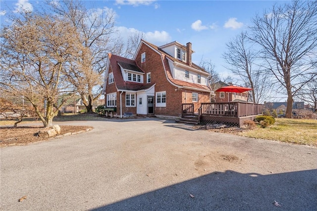 view of front of house with brick siding, a wooden deck, driveway, and a chimney