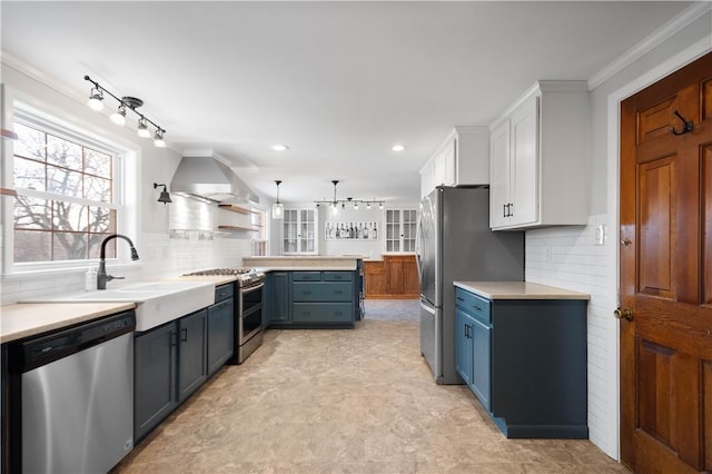 kitchen featuring wall chimney range hood, ornamental molding, stainless steel appliances, white cabinetry, and blue cabinets