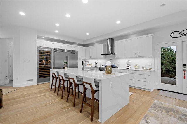 kitchen featuring a breakfast bar area, light countertops, built in refrigerator, wall chimney exhaust hood, and light wood-type flooring