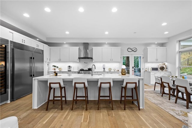 kitchen featuring light wood-style flooring, wall chimney range hood, light countertops, and stainless steel built in fridge