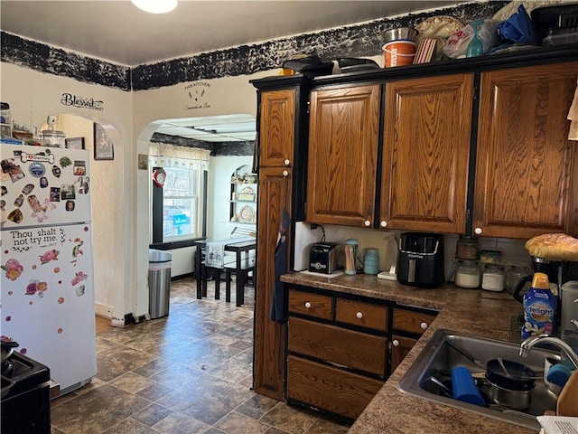 kitchen with arched walkways, freestanding refrigerator, a sink, stone finish floor, and brown cabinets