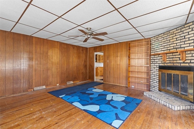 unfurnished living room with a brick fireplace, hardwood / wood-style floors, a ceiling fan, and wood walls