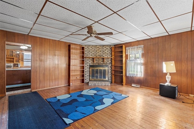 unfurnished living room with a brick fireplace, a ceiling fan, wood walls, and wood-type flooring