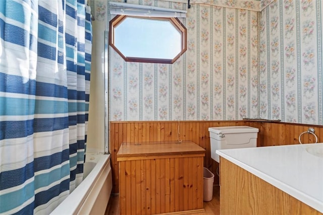 bathroom with wallpapered walls, shower / tub combo, toilet, and a wainscoted wall