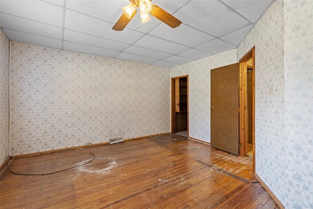 empty room with wallpapered walls, baseboards, a drop ceiling, hardwood / wood-style flooring, and a ceiling fan