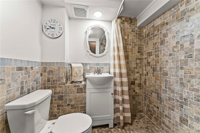 full bathroom with visible vents, a wainscoted wall, toilet, a tile shower, and tile walls