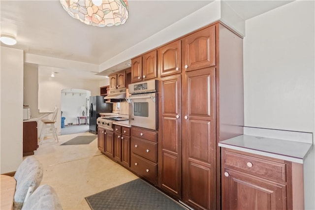 kitchen with brown cabinetry, freestanding refrigerator, cooktop, under cabinet range hood, and stainless steel oven