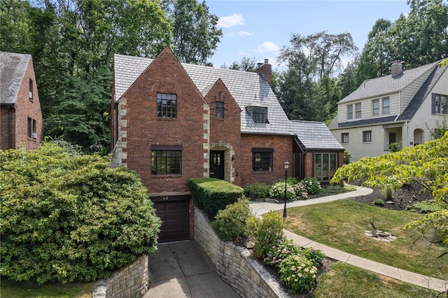 tudor-style house with driveway, a high end roof, an attached garage, a chimney, and brick siding