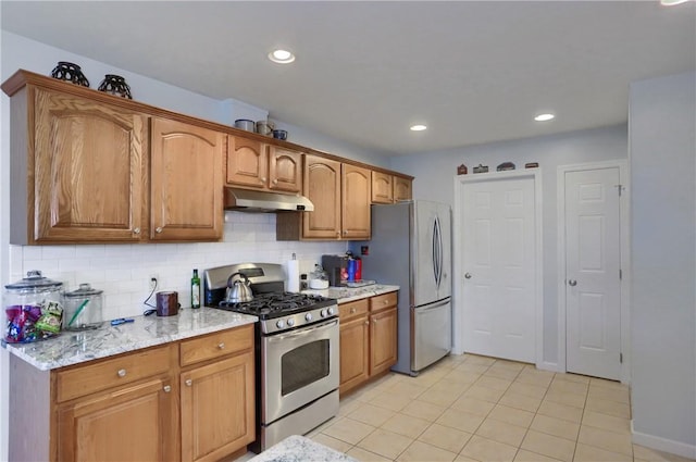 kitchen with under cabinet range hood, light stone counters, backsplash, stainless steel appliances, and brown cabinetry