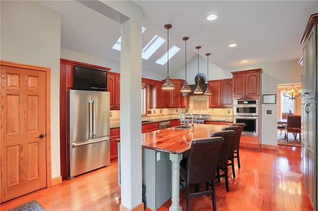 kitchen with vaulted ceiling with skylight, a kitchen island with sink, light wood-style floors, appliances with stainless steel finishes, and backsplash