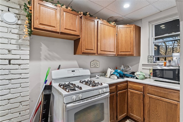 kitchen featuring brick wall, a drop ceiling, light countertops, white gas range oven, and stainless steel microwave