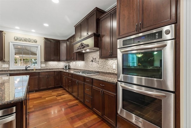 kitchen featuring wood finished floors, dark stone counters, dark brown cabinets, appliances with stainless steel finishes, and tasteful backsplash