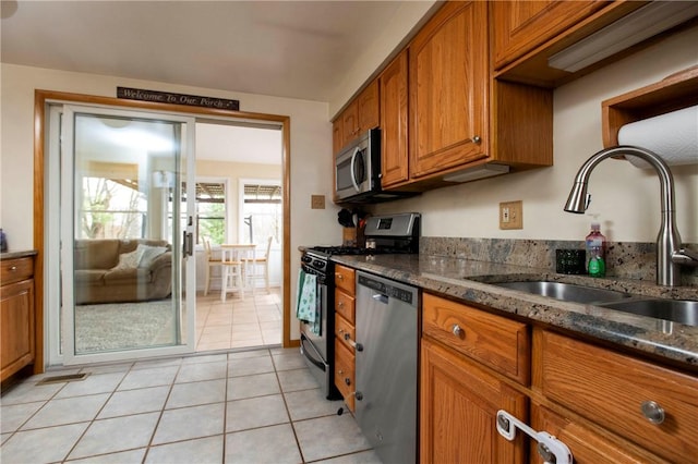 kitchen featuring light tile patterned floors, dark stone countertops, brown cabinets, stainless steel appliances, and a sink