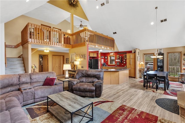 living room featuring stairs, visible vents, high vaulted ceiling, light wood finished floors, and ceiling fan with notable chandelier